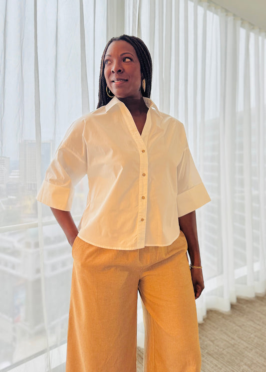 Woman wearing a white shirt and pants standing in front of sheer curtains.