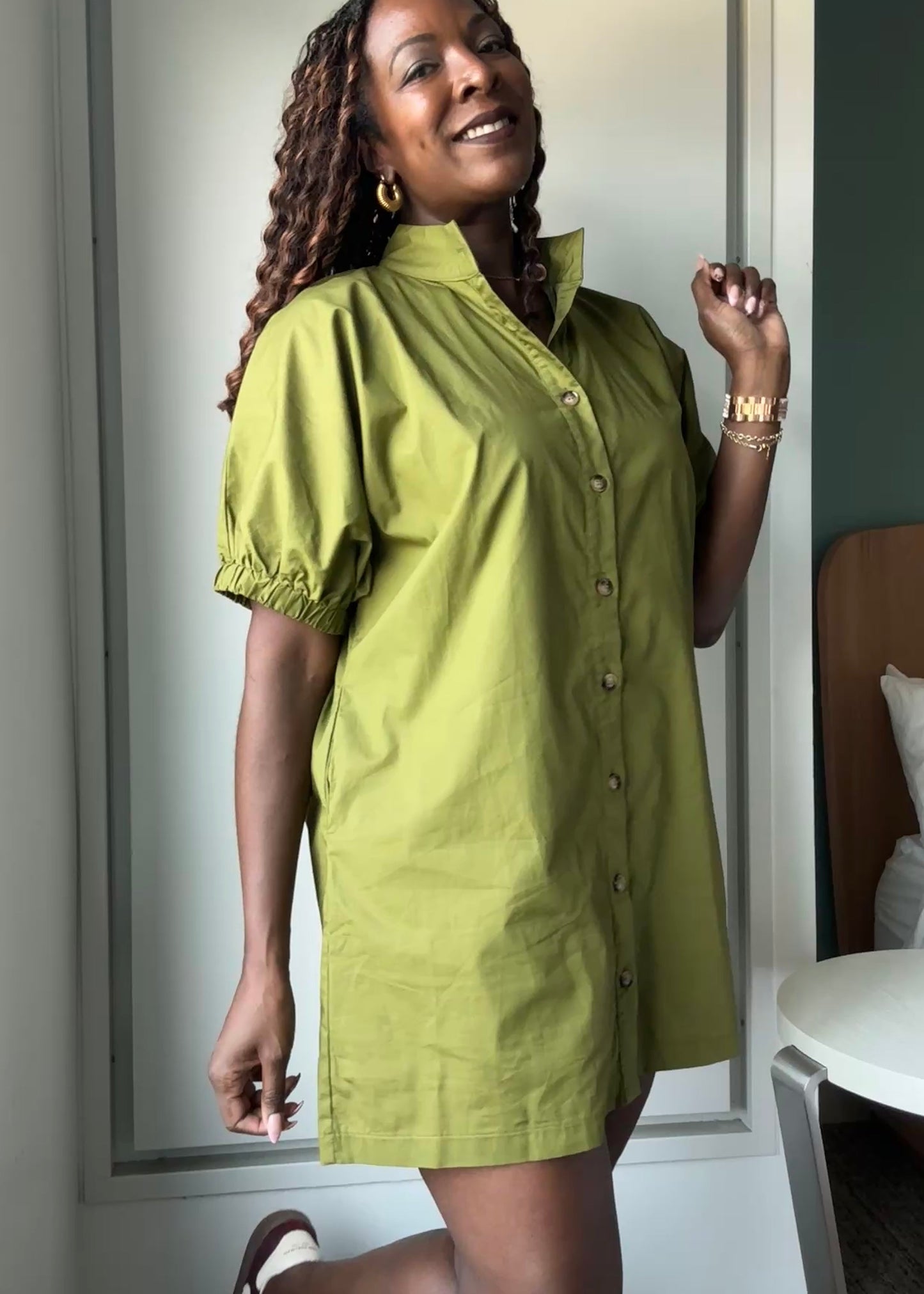 A woman wearing a short-sleeve button-front shirt dress in olive green, standing indoors near a window and bed.