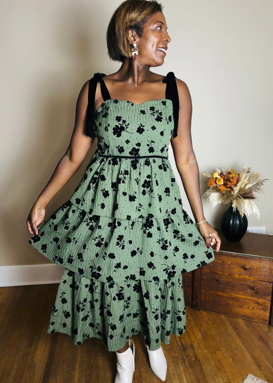 Woman wearing a green floral dress in a room with wooden floor and wall.