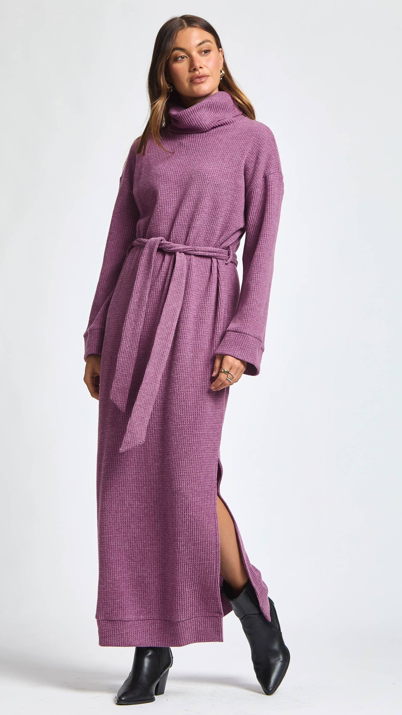 Model wearing a brushed thermal maxi dress with a relaxed cowl neckline, removable waist tie, and side slits. Soft, textured knit fabric drapes gently through the body with a long, elegant silhouette. Sleeveless design with a cozy, elevated feel