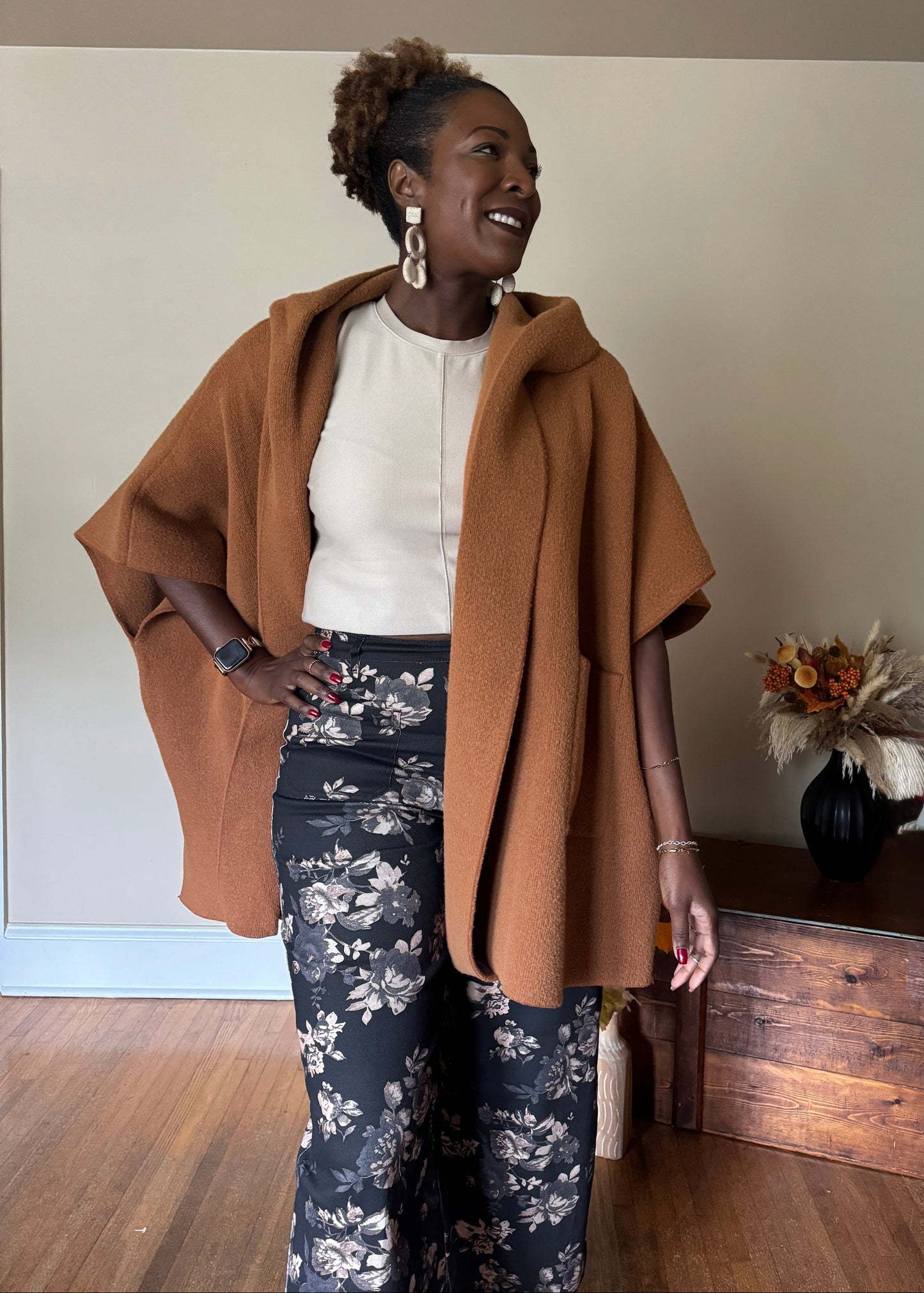 Woman wearing a brown coat over a white top and floral pants, standing indoors.