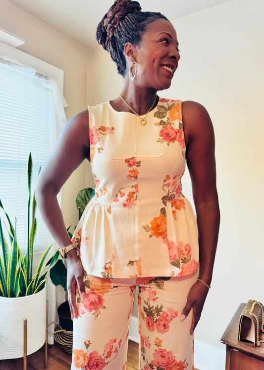 Woman wearing a floral peplum sleeveless top with matching pants standing in a room with plants and a window.
