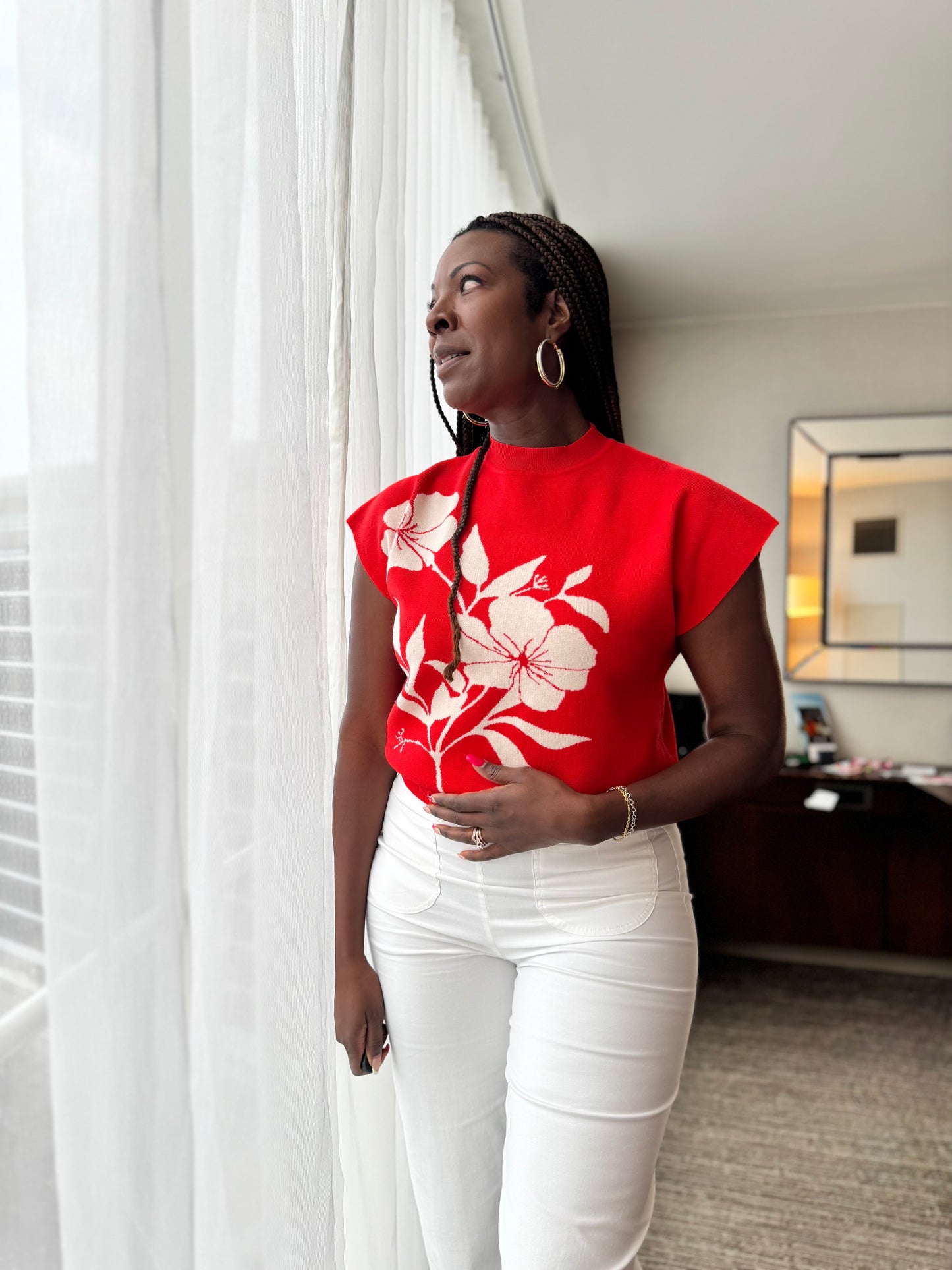 Woman wearing a red floral top and white pants standing in a room with a window and desk in the background.