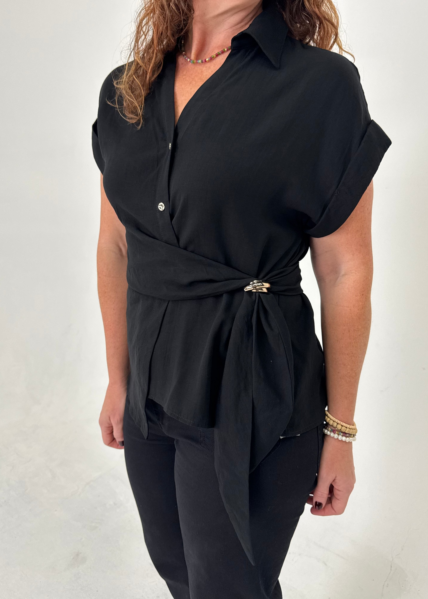 A woman wearing a black short-sleeve collared blouse with a wrap-style waist and a small gold-toned belt buckle, paired with black pants, against a plain light background.
