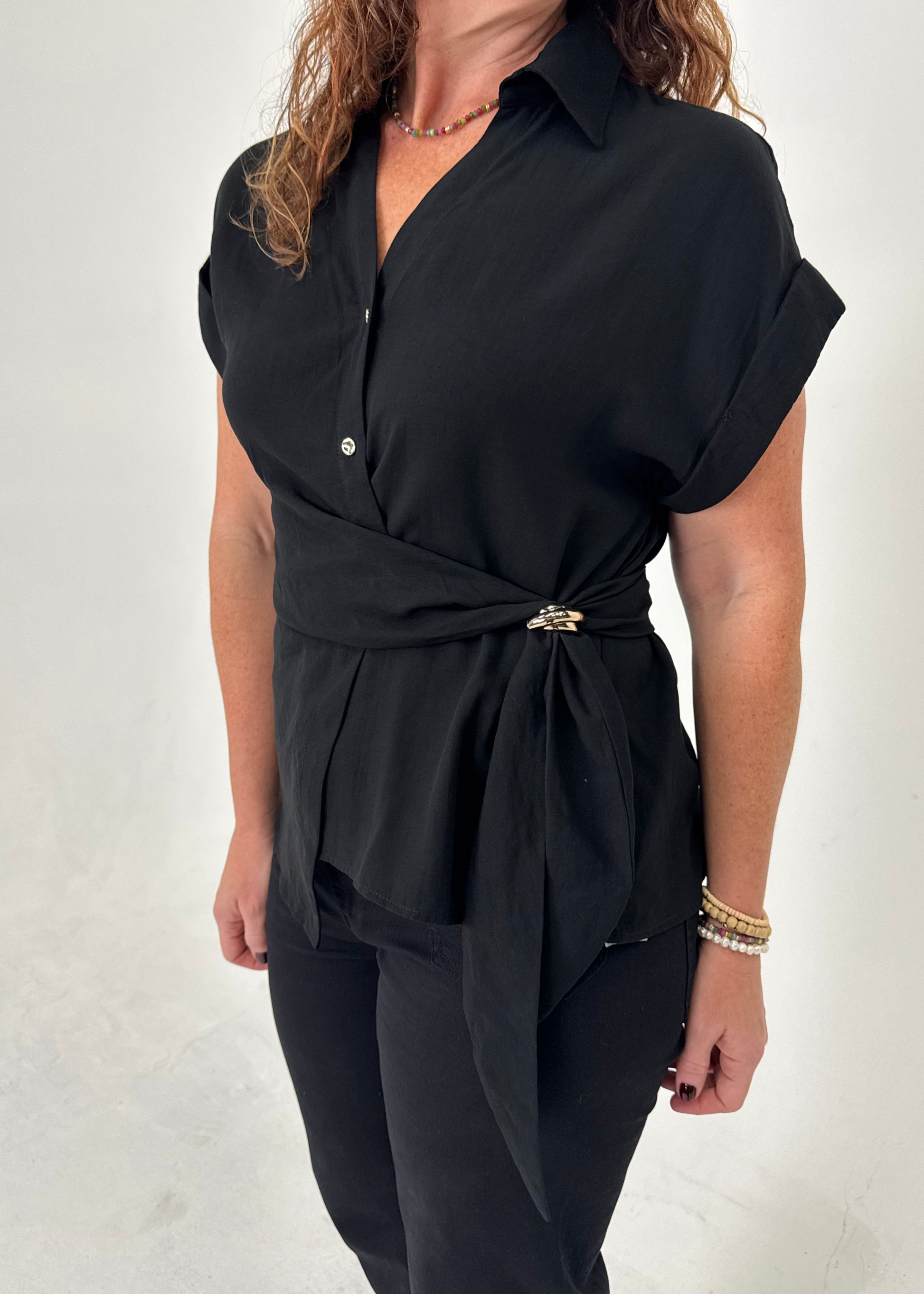 A woman wearing a black short-sleeve collared blouse with a wrap-style waist and a small gold-toned belt buckle, paired with black pants, against a plain light background.