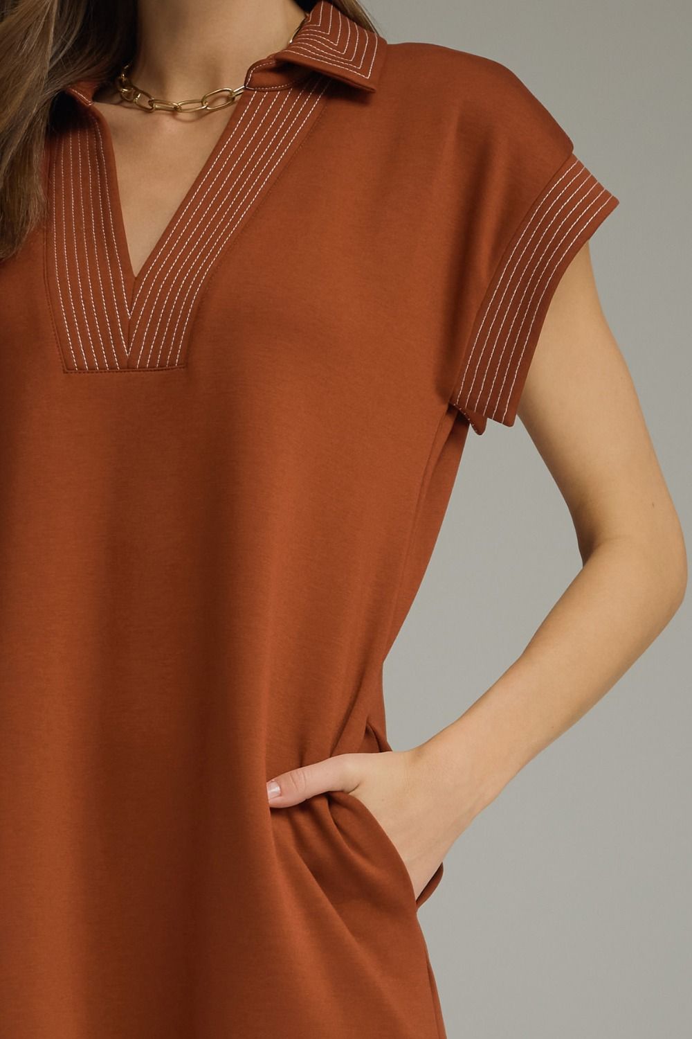 A close-up of a woman wearing a rust-orange short-sleeve collared dress or tunic with contrasting striped trim along the collar and sleeve edges, paired with a gold chain necklace, against a solid gray background.