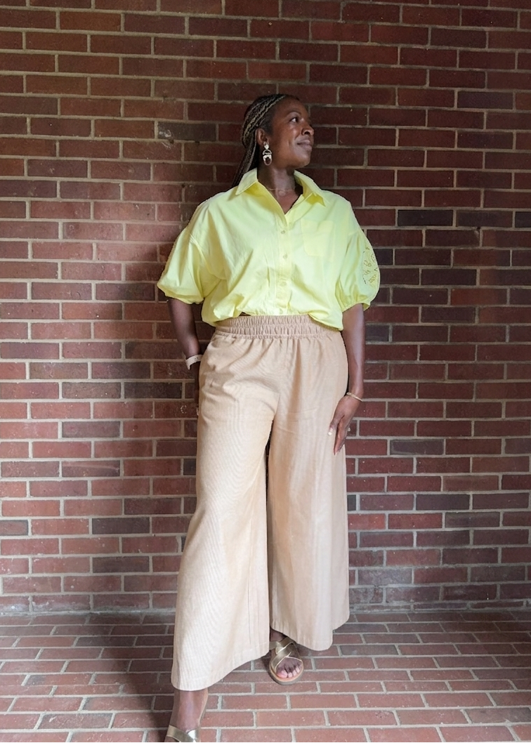 Woman in a yellow shirt and wide linen pants standing against a brick wall.