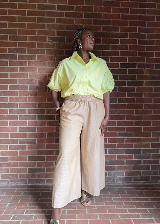 Woman in a yellow shirt and wide linen pants standing against a brick wall.